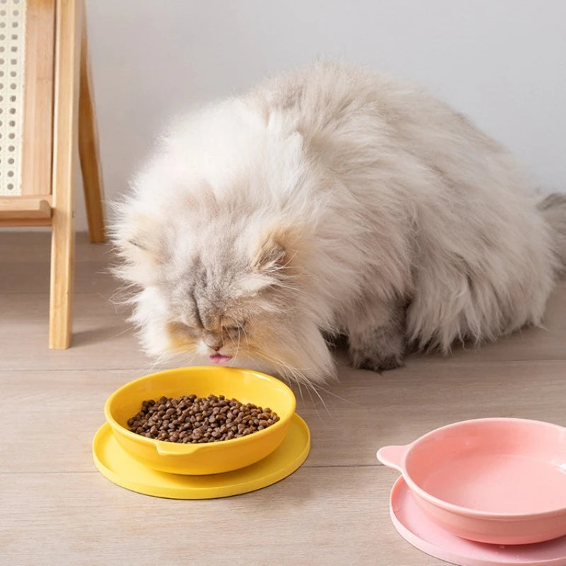 Cat eating from a yellow bowl on a wooden floor with pink bowls in the background.