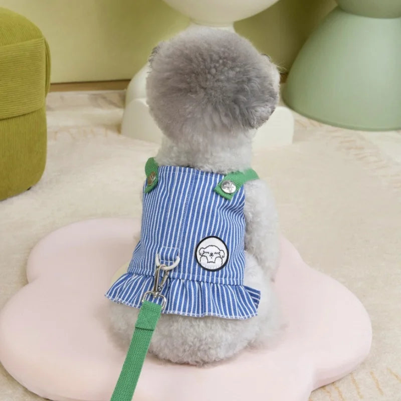 Small dog wearing a blue striped dress with green straps on a pink heart-shaped cushion.