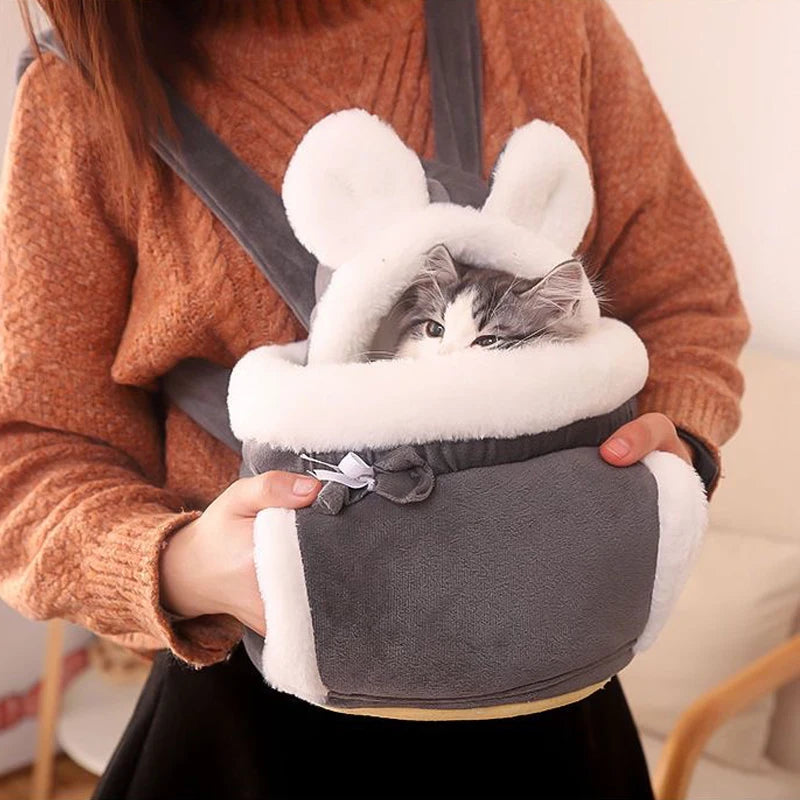 Cat in a gray and white bunny-style carrier held by a person wearing an orange sweater.