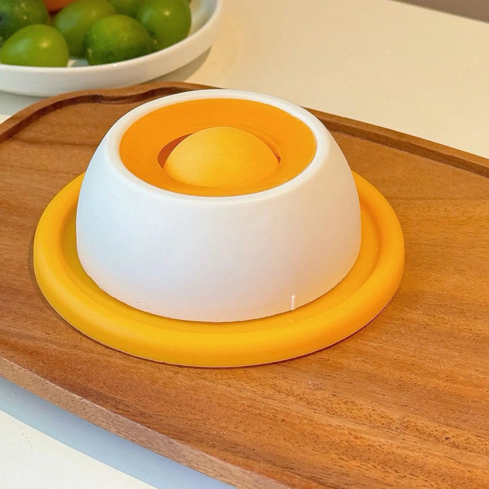 White and orange kitchen tool on a wooden surface with green fruits in the background