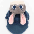 Plush toy resembling a gray creature with large eyes and pink ears, wrapped in a blue blanket on a white background.